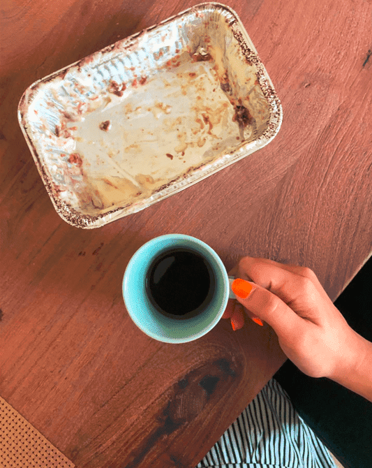 A hand holds a cup with coffee in it over a wooden table. There’s an empty aluminium food container with traces of cream and chocolate on the table next to the cup. 