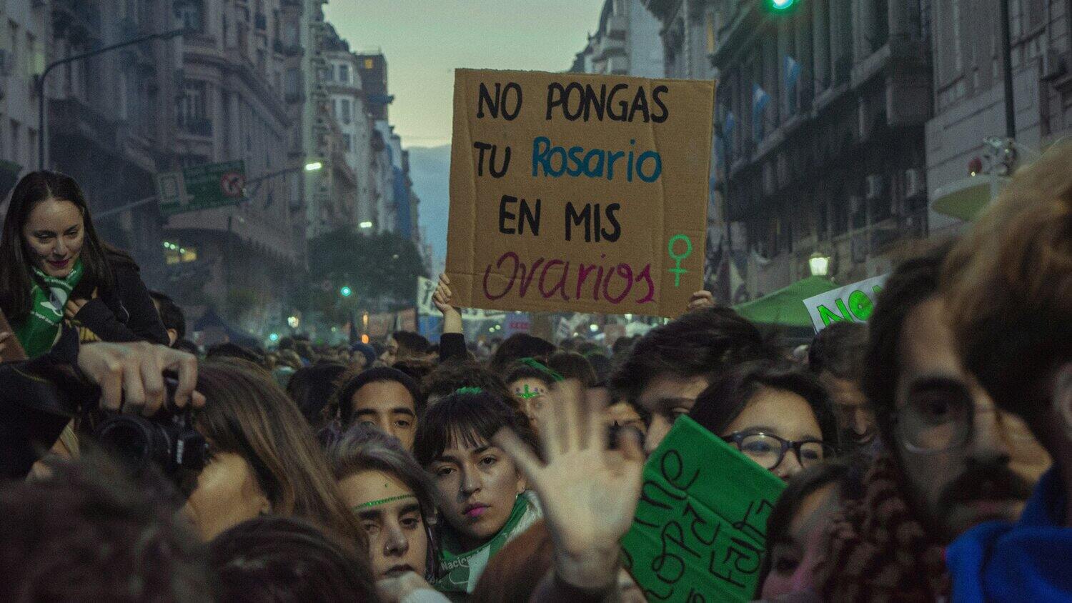 Image of hundreds people on the street at night. Many wear green headbands and are facing forward. A few people at the front of the crowd turn back, and others in the middle of the crowd are holding up a sign.