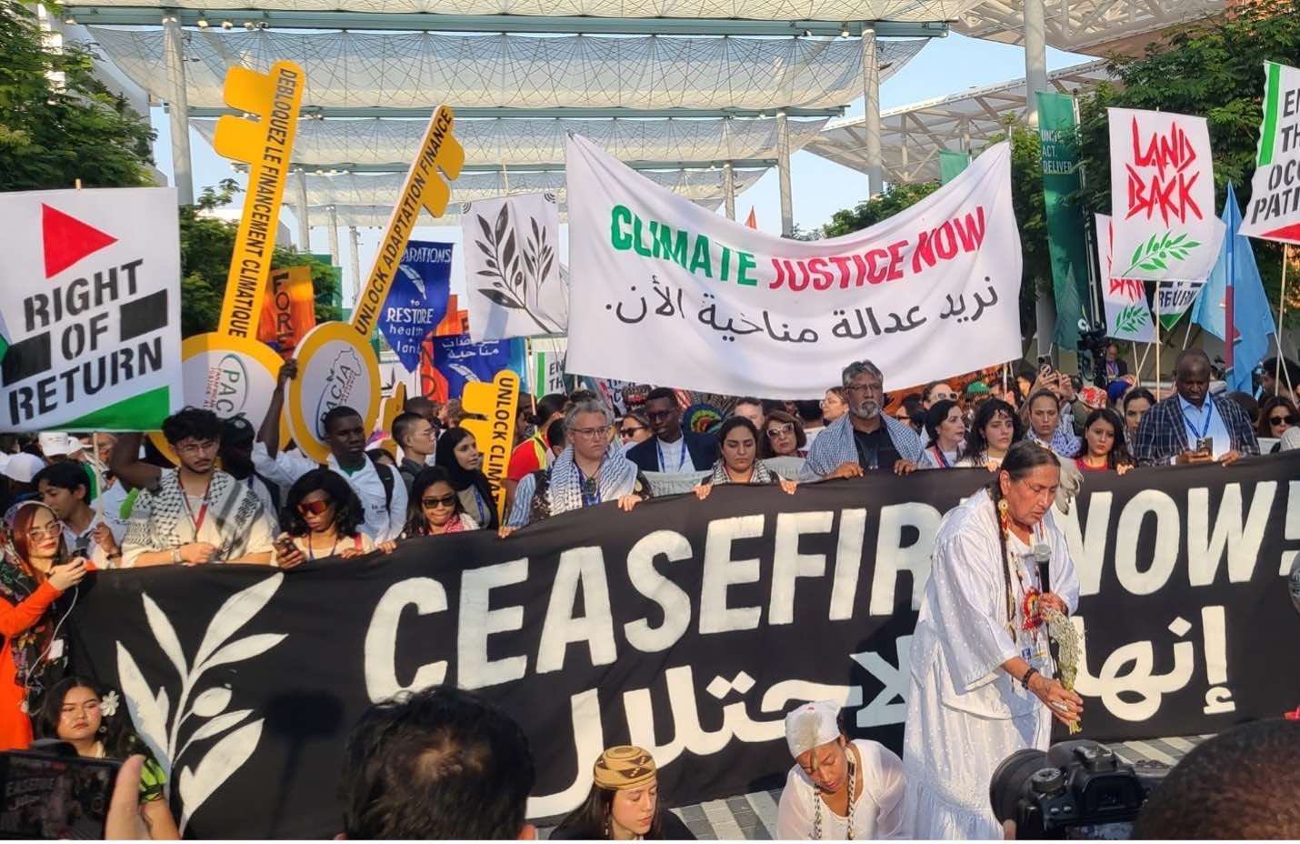 Rows of men and women stand closely, holding signs including "Right of Return" and "Climate Justice Now" in green, red, and black lettering. The group stands behind a black horizontal sign that reads "Ceasefire Now!" in English and Arabic. A woman in white with a plant in one hand and a microphone in the other stands in front of the sign.