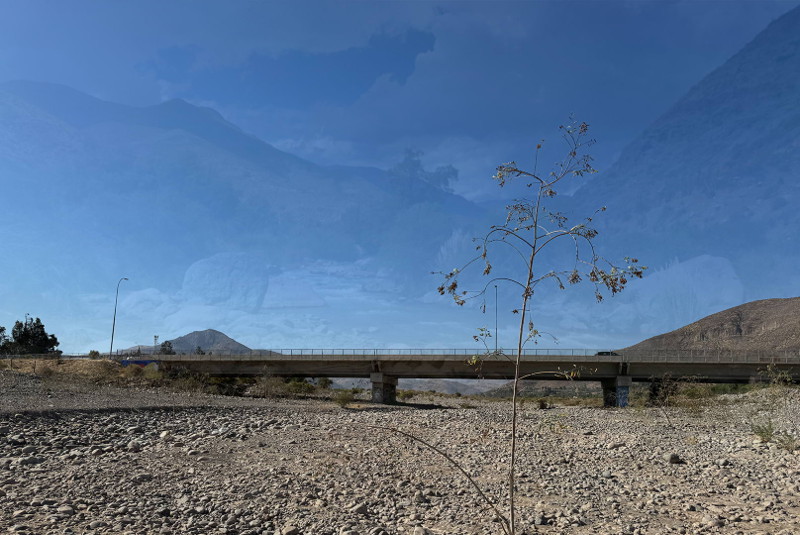 An image showing a rocky, grey-brown, dried river bed and a bridge that crosses over. The top half of the image shows a clear blue sky.