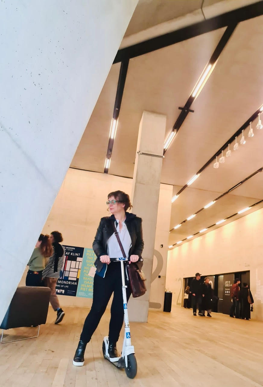 A white, middle-aged woman stands with one foot on a scooter painter white with disability stickers. She is inside a museum with a bright, modern aesthetic.