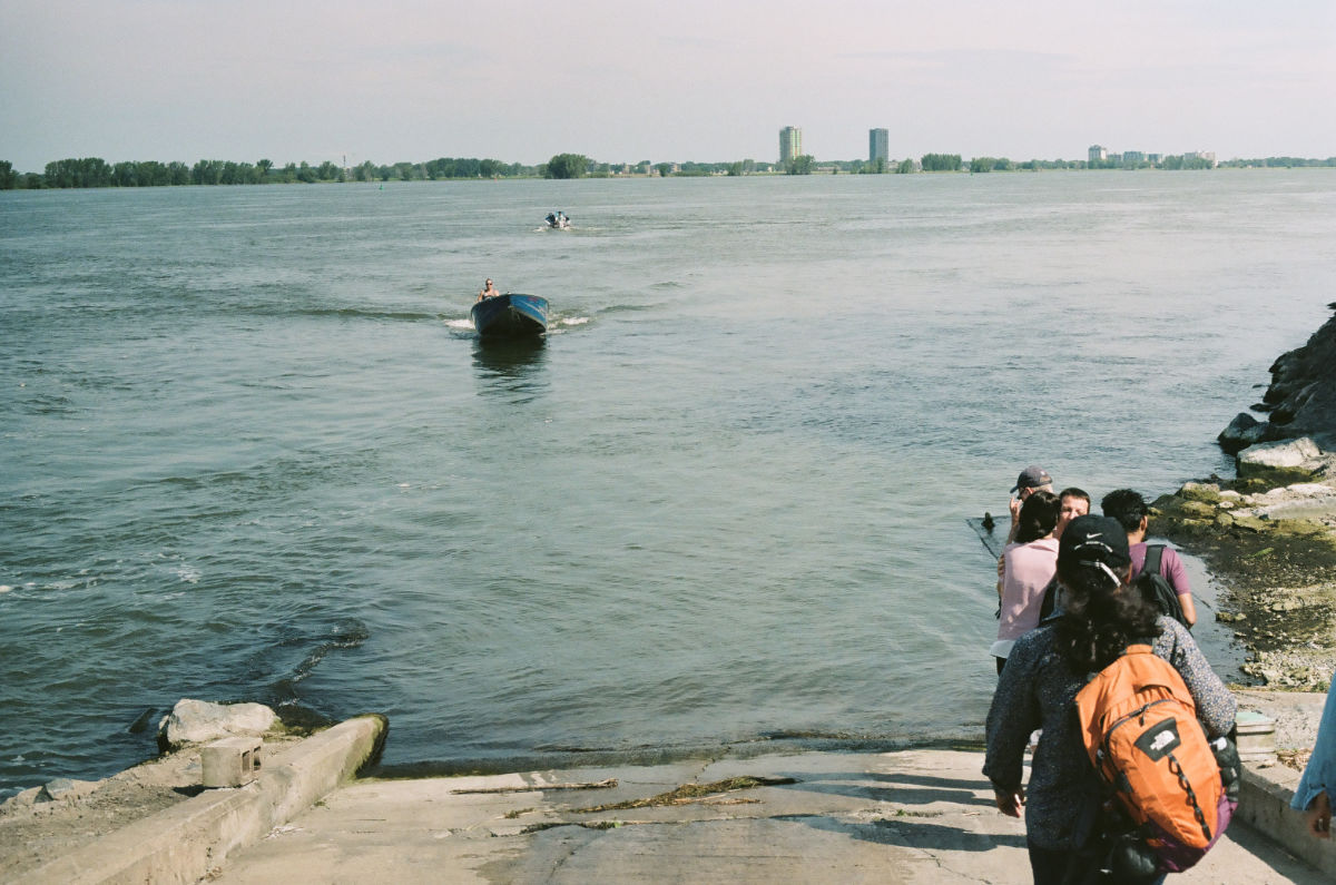 A river with a person in a small boat appears to be moving toward students on a concrete boat launch in the foreground; there are buildings and trees on the other side of the river.