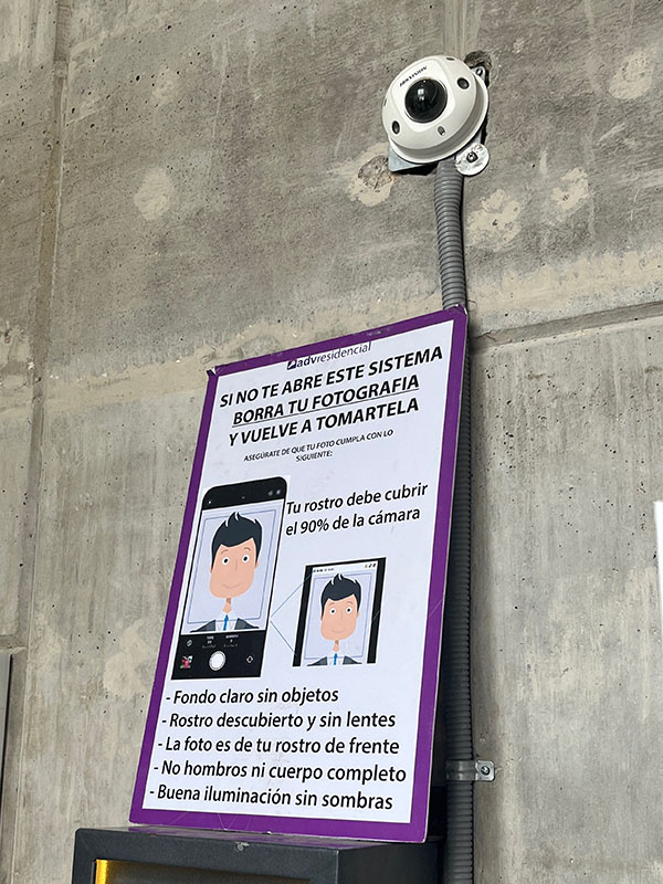 A booth with a camera and a big sign against a wall. The sign has instructions on what to do to have one’s photograph taken. The room where the booth is looks to have unfinished concrete walls, a rough presentation for an entrance to a luxury gated community.