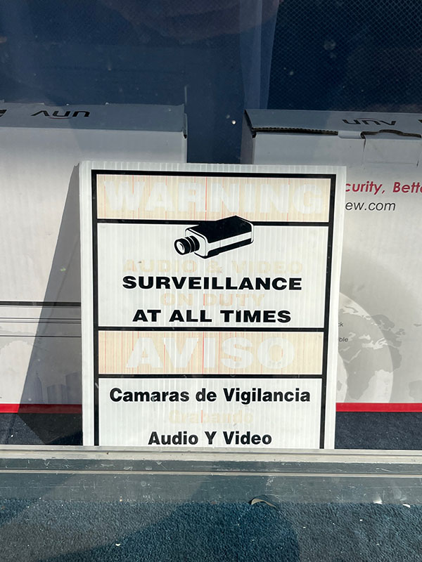 A faded foam board sign is propped against an empty box on a storefront window. The sign combines signs in English and Spanish: “Warning / aviso,” “Surveillance at all times,” “Surveillance cameras, audio and video.”