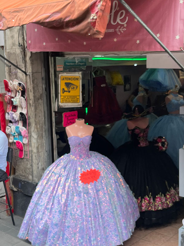 Storefront of a quinceañera dress shop has several sequined A-line ball gown silhouette dresses in bold colors. There is a sign with a camera that reads, “Attention! For your security, this place operates CCTV cameras.”