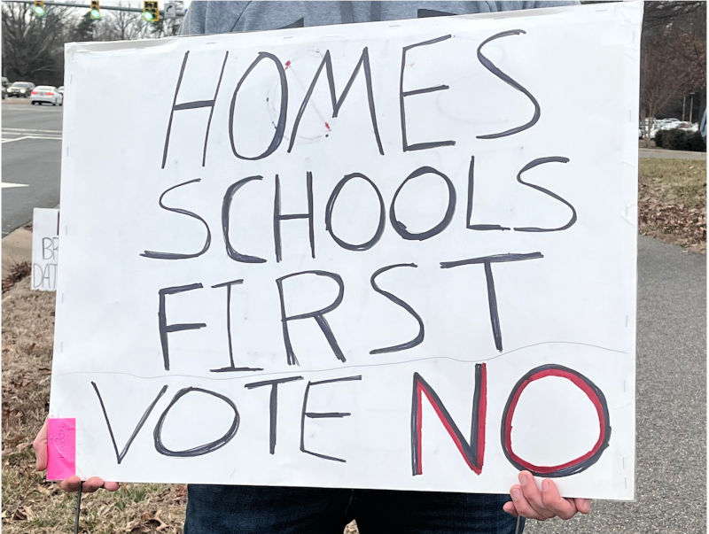 Person holding handmade sign that reads "Homes Schools First Vote No."