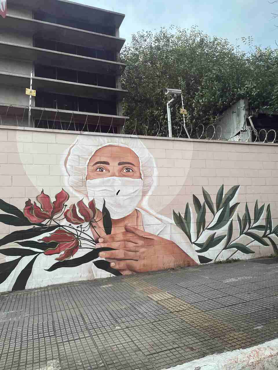 A brick wall with a mural depicting a person with a surgical cap and gown holding the stems of a plant, which has narrow leaves and red flowers.