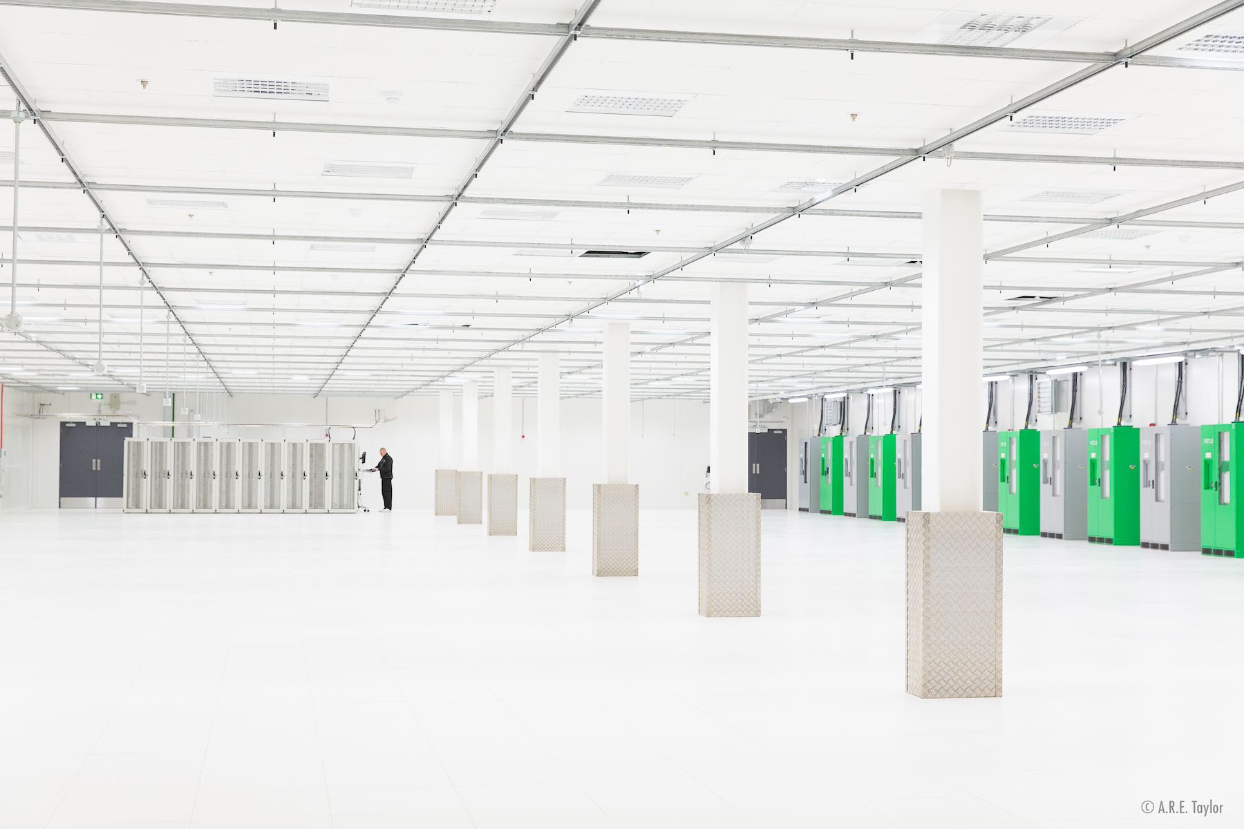 An image of a large white room containing several painted white and concrete pillars. At the back of the room stands a man in black surveying a row of grey data servers.