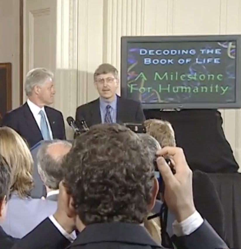 President Bill Clinton and Dr. Francis Collins stand at a podium in the background, next to a digital screen which reads the following text in title case: "Decoding the book of life: A milestone for humanity." In the foreground, the back of a photographer's head can be seen as he takes a picture of the scene.