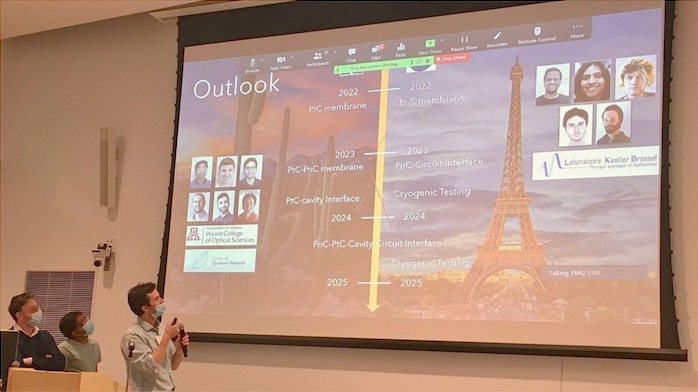 Two people wearing face masks stand in front of a slideshow on a projector screen; the slide shows pictures of cactuses and the Eiffel Tower, and reads "Outlook."