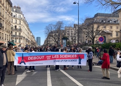 A crowd of people marches past stately multi-story buildings, holding a blue, red, and white banner reading "Debout pour les sciences."