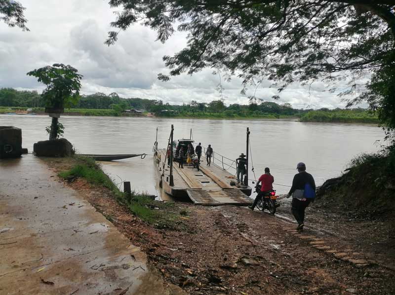In the image, a modern flatboat is seen in the center, carrying four people, a car, and a motorcycle. Additionally, two more people are boarding from the riverbank. In the background, a tropical forest typical of Colombia can be seen.