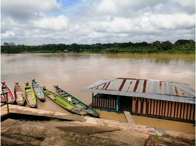 In the image, there is a house built within the river on wooden stilts, connected to the shore by a bridge. Several canoes are seen beside it. A wide river runs through the center of the image, and on the horizon, a thin line of typically Colombian forest can be seen.