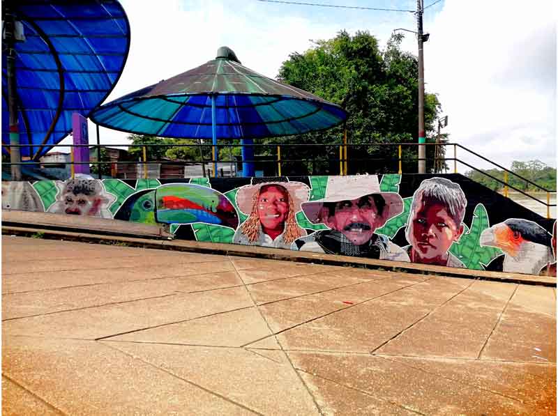 The image shows a graffiti mural on a street wall. At the top of the image, a large umbrella for sun protection and the sky can be seen. The graffiti features a series of faces representing the diversity of the local population. In the center, there is a middle-aged man with a mustache; to the left, a boy with features next to a bird. On the right, there is an teenage girl, a pelican, and a titi monkey.