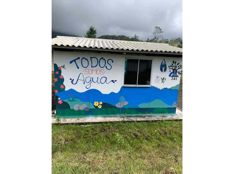 The image shows a small house surrounded by grass. The house is blue on the lower part and white on the upper part, decorated with drawings of flowers and stones simulating a river. On the right side, the phrase "Todos somos agua" ("We are all water") is written. In the center, there is a window, and on the left side, a drawing of a raised hand, a tree with a seated person, and below it, the initials "ZRC".