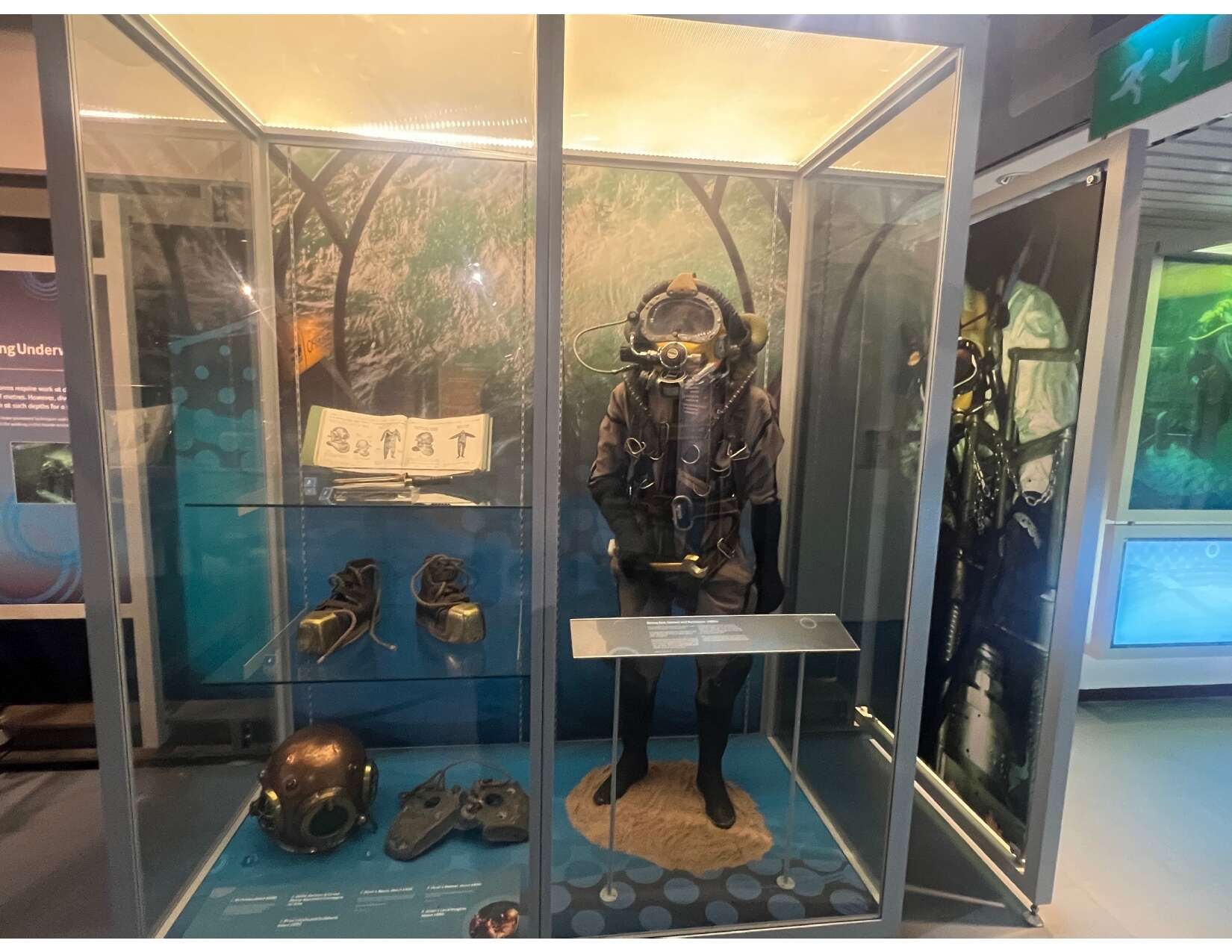 The image shows a museum display featuring an early model of a diving suit, diving shoes, weights, and a helmet. Additionally, an old book is exhibited, displaying sketches of diving suits and helmets.