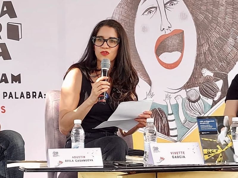 Vivette García Deister presents a book while holding a microphone in one hand and a sheet of paper in the other. 