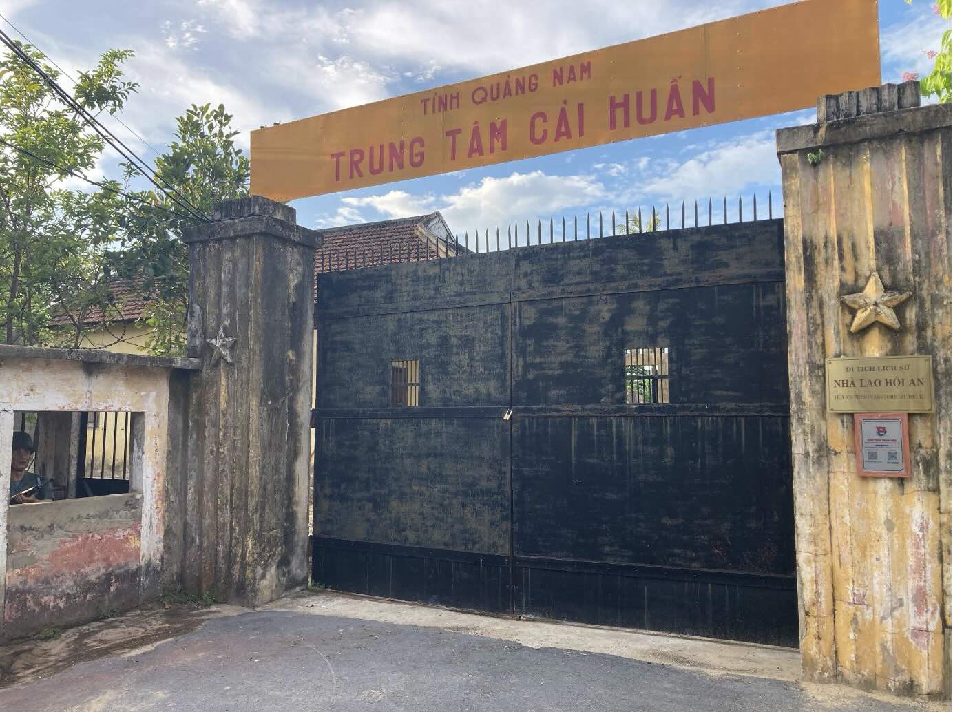 A large, closed iron gate blocks off a courtyard for the viewer. An orange sign above it in red letters reads, in Vietnamese "Tinh Quáng-Nam Trung Tám Cái Huán" ("Quang Nam Province: Correctional Center")