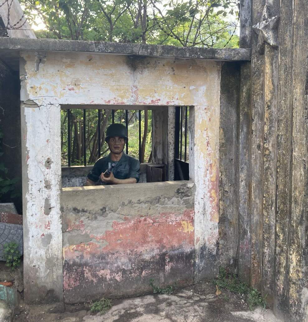 A lifelike statue of a Vietnamese soldier in combat fatigues kneels behind a small bunker and aims a fake rifle at the viewer in a threatening manner.