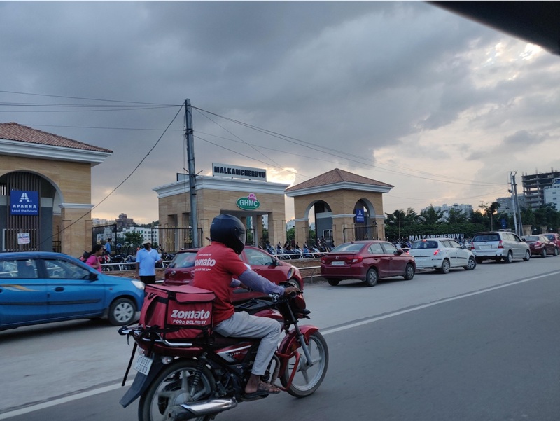 A delivery person dressed in a red uniform (of the company Zomato) can be seen riding on his bike past a prominent public spot (the Malkacheruvu) in the city, managed by the municipal corporation body GHMC, on a cloudy day. Cars are parked outside, and a few people can be seen in the background.