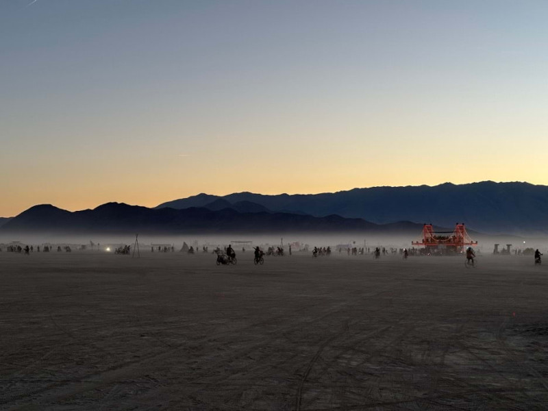 A desert landscape at dusk. In the distance are the silhouettes of people and an electric sculpture of the Golden Gate Bridge.