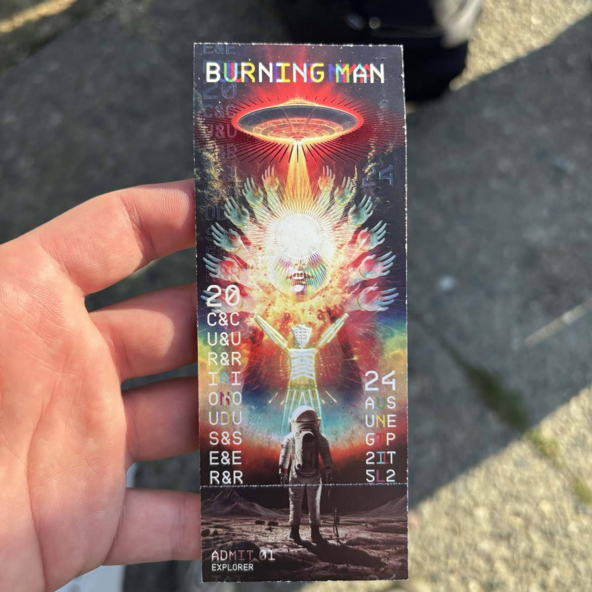 A hand holding a paper ticket, depicting a stylistic UFO and an astronaut. It displays the title Burning Man as well as event dates.