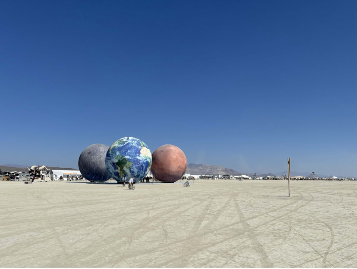 A desert landscape during the day. In the distance are the silhouettes of people and three large globes, one of which is the earth.