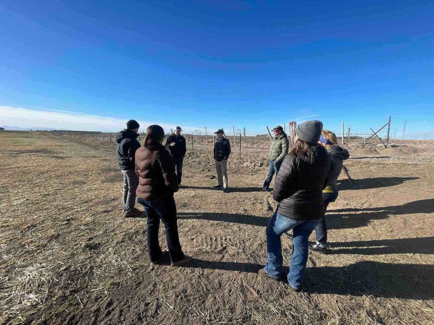 Eight adults stand in a circle on a field with brown soil visible underfoot.