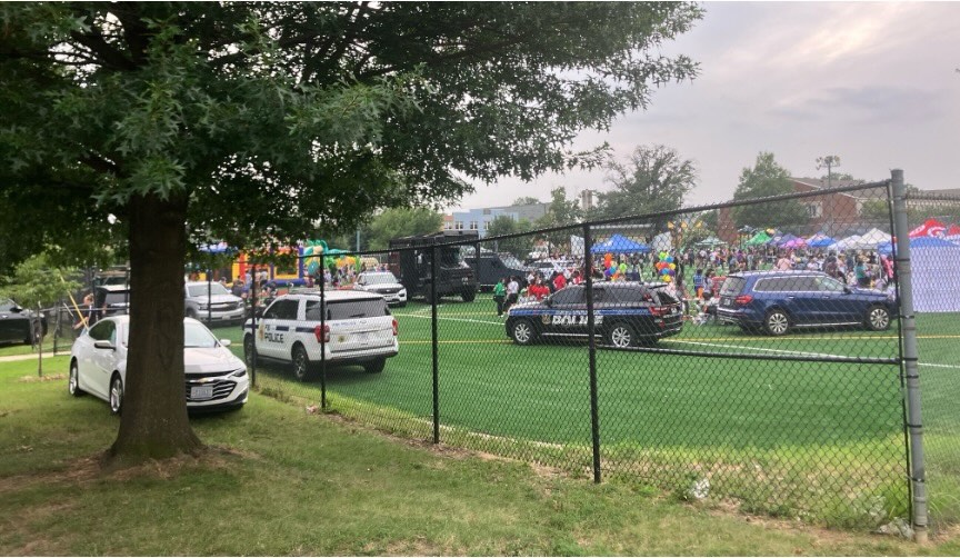 A photograph of a summer festival in a grassy field, with a black chainlink fence and several parked police cars between it and the viewer. 