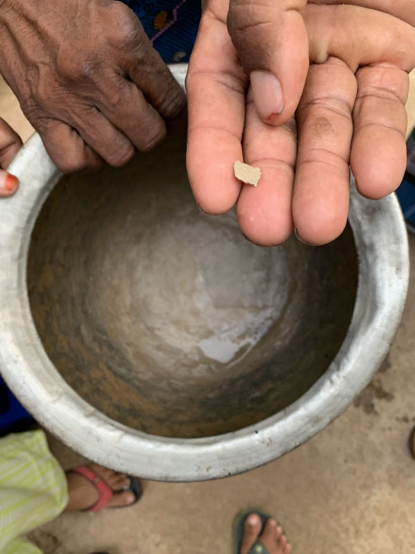 The participant is showing a pale brownish piece of chemical residue on the palm of the participant. The participant's thump nail is chipped at the edge, exposing the wounded skin. Beneath the participant's hand is a man's hand holding the aluminum pot used for heating water. There is no water in the pot and the bottom of the pot has a layer of chemicals that have saturated along it surface while heating. 