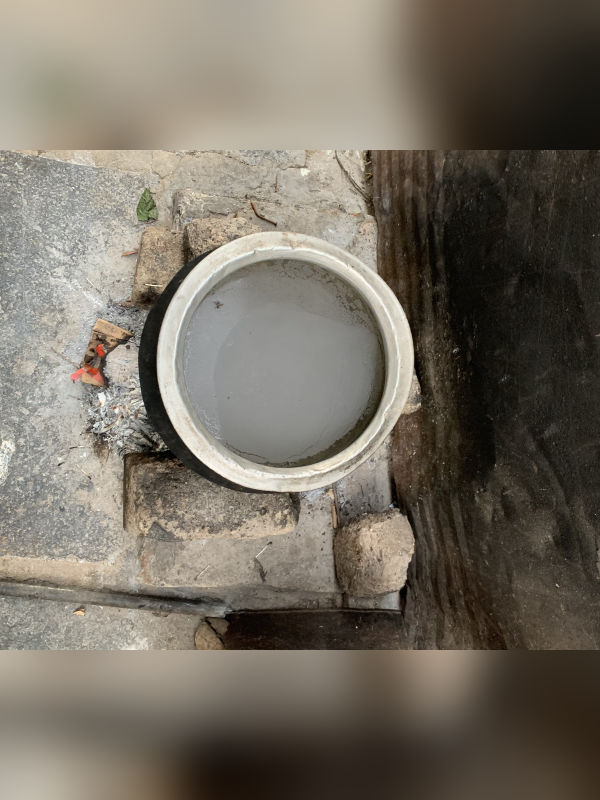A aluminum pot with water to the brim captured from above. The pot is placed on bricks on either edges. In between the bricks are small pieces of wood set on fire and its residual ash. A mild flame below the pot is visible on the image. The warm water in the pot has a white layer of chemicals that has risen to the surface during boiling.