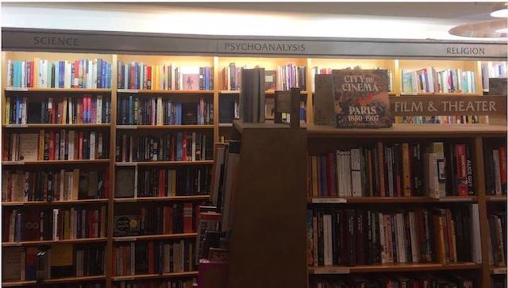 Shelves full of books in a NYC bookstore, with signs atop designating the contents of the bookshelves. To the left “science”, in the middle “psychoanalysis”, on the right “religion”.
