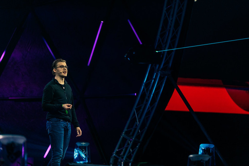 A white man in jeans and a black jacket stands on stage, speaking into a headset microphone. 