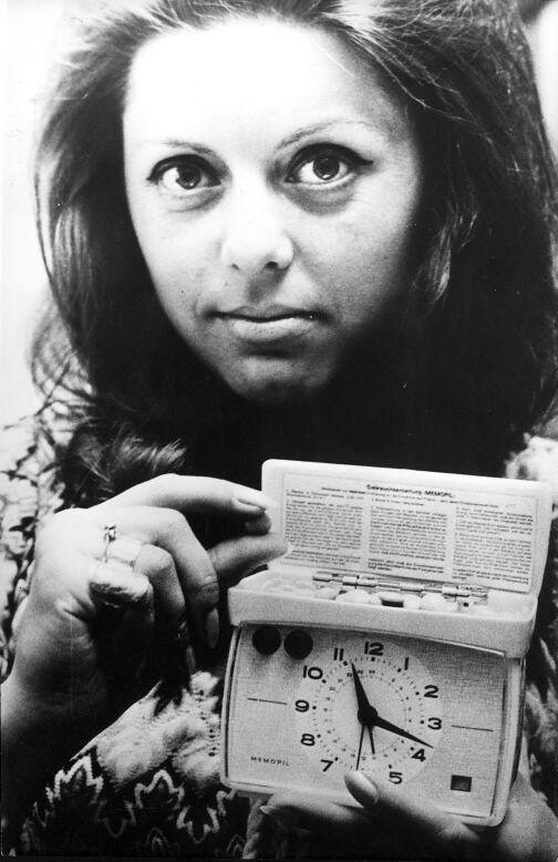 A black-and-white photograph of a white woman in her twenties or thirties. She is gazing directly at the camera and holding up an alarm clock with a pill tray attached to the top, allowing for reminders to take birth control. In her other hand, which has a ring, she is holding a single pill.