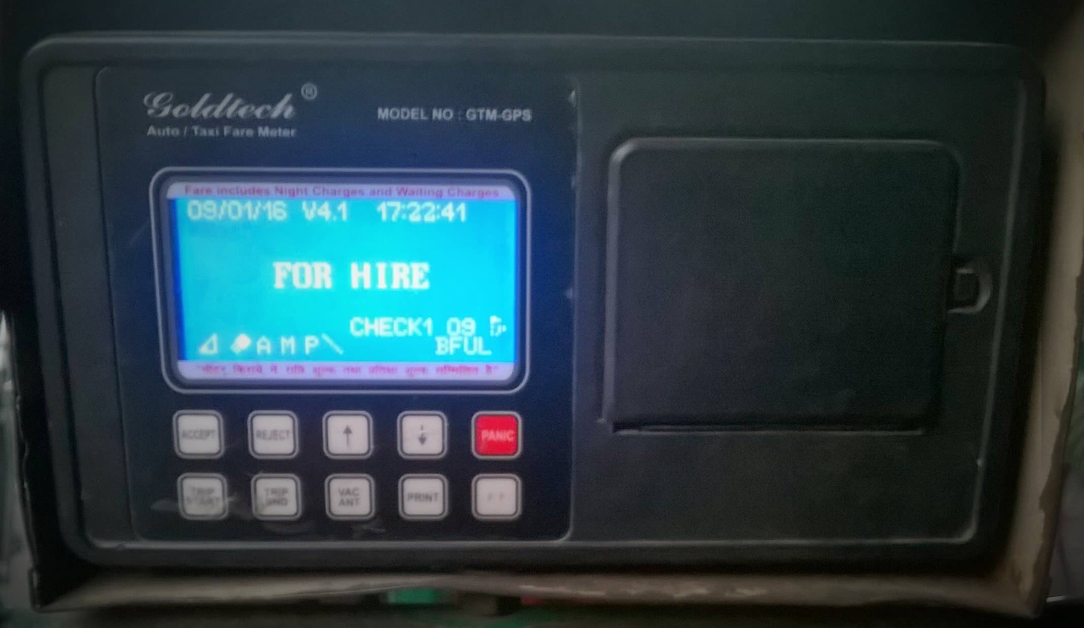 A color photograph of a somewhat small electronic box. The right half of the box is smooth and blank while the left side has a glowing blue digital screen. Below the screen there are 9 grey buttons and 1 red button that says "PANIC." The screen displays a date, time, and the phrase "FOR HIRE."