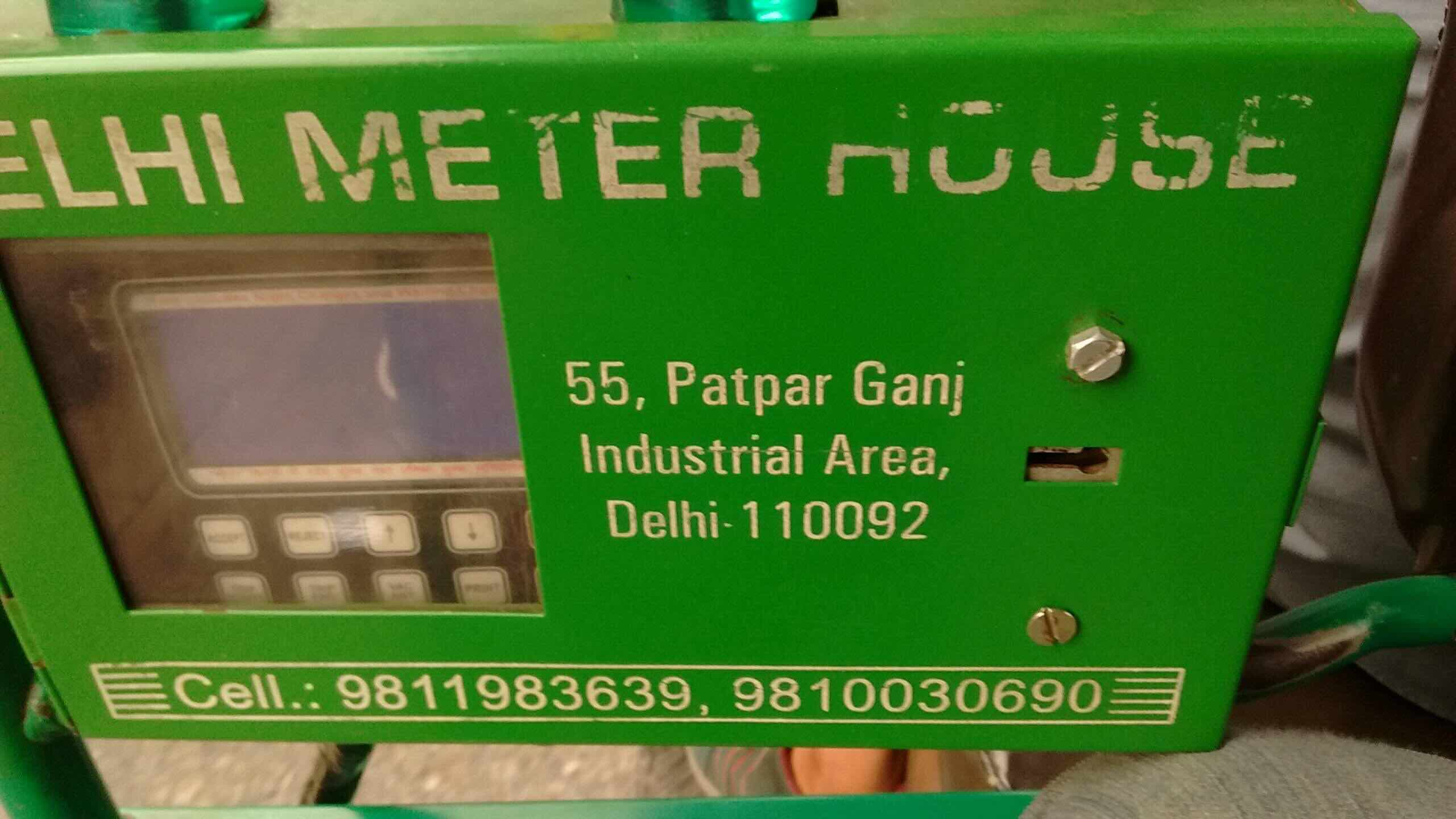A color photograph depicting the same device from image 1, this time encloseed in a green metal box. The box reads DELHI METER HOUSE on top with an address in the middle and phone numbers at the bottom. On the left, a smudged window affords a view of the screen of the device.