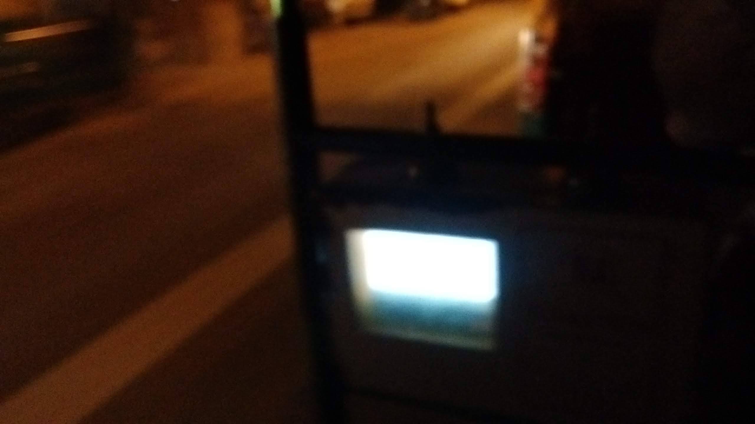 A blurry photograph from the inside of an autorickshaw at night moving down the street. The bright white screen of the meter takes up the center of the frame and beyond it is the obscured figure of the driver.