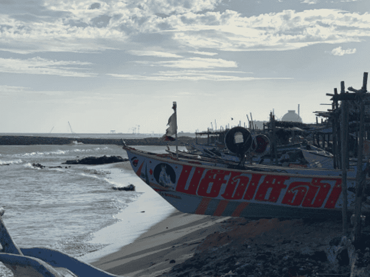 A view of Kudankulam's sea shore. In the foreground of the image, waves touch the shore where traditional wooden boats, painted in red and blue, are moored on beach sand. In the background, is the tomb of KKNNP's nuclear containment.