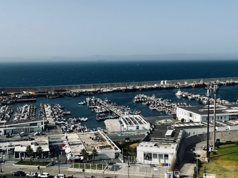 Elevated view of the Tanger-Ville fishing port. Port facilities and docks are enclosed by a sea wall. Beyond is a container ship sailing through the Strait of Gibraltar. The Spanish coast is visible in the distance. Photo taken by the author.