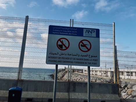 The perimeter fence of the Tanger-Ville port. On the fence is a sign reading “fishing and swimming forbiden [sic] in the port” in Arabic, French, and English. 