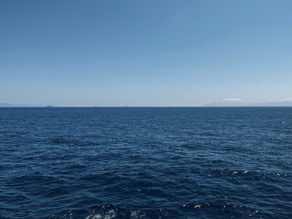 A horizon, sea-level view of the Strait of Gibraltar from the deck of a fishing boat. Small shapes of container ships dot the horizon.