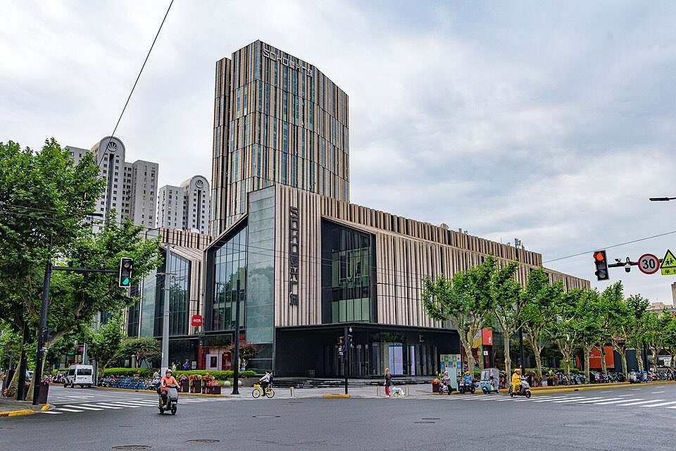 A color photograph of an urban landscape with low, flat, sleek and modern building in the foreground and a taller tower behind it in the same style. Motorists on scooters can be seen on the streets intersecting in front of the building.