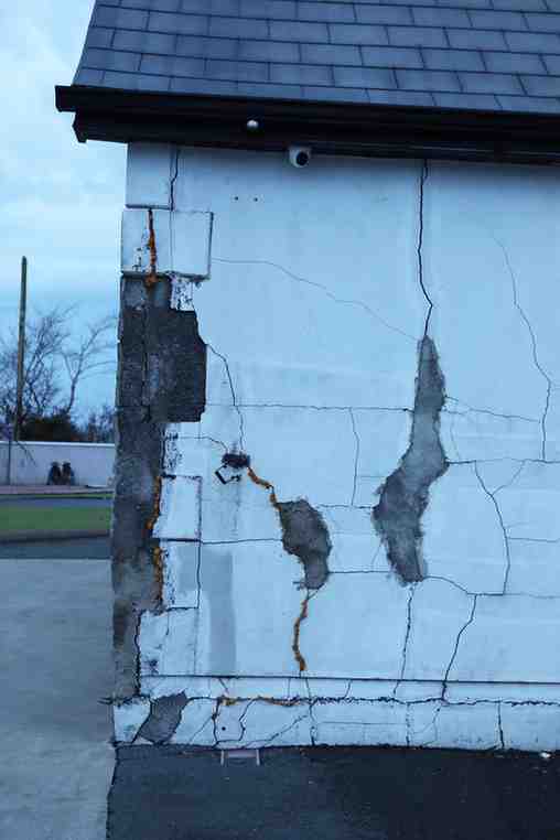 Photograph of a building corner showing extensive wall damage with large cracks and exposed underlying material. The white exterior wall has multiple vertical and horizontal fractures, with visible rust stains and deterioration near the base. The image shows blue and grey skies and the wet concrete indicates it had previously been raining.