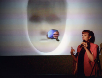 Artist speaking into a microphone in front of a projected blurred digital face during a presentation.