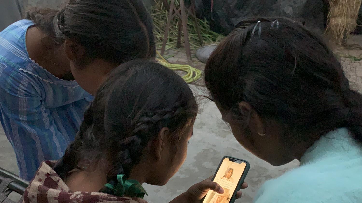Three young girls in a rural setting gather around a smartphone, closely watching content on the screen together.