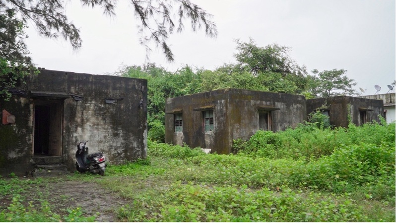 Abandoned concrete buildings overgrown with vegetation, with a motorcycle parked near the entrance of one structure. The weathered, moss-covered walls and surrounding dense greenery suggest long-term neglect.