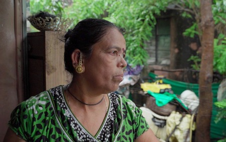 Kalpana, a resident of Popharan, seated and gazing into the distance. She wears a green patterned top and gold earrings. A lush green outdoor area with household items is visible in the background.