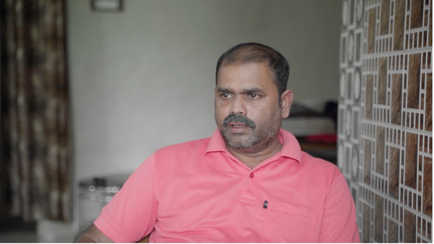 Harkesh Tamhore, a contract worker at the Tarapur Power Plant, seated indoors and looking to the side with a serious expression. He wears a pink polo shirt. A textured wall and soft background interior are visible behind him.