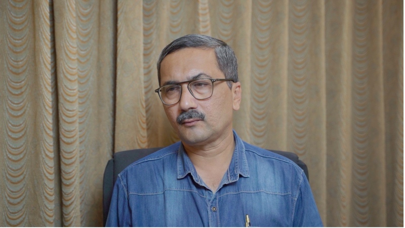 Neeraj Raut, a journalist, seated in an office chair and looking directly at the camera with a composed expression. He wears a denim shirt and glasses. A beige curtain forms the background.