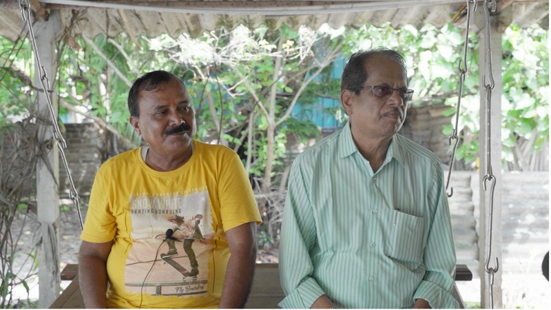 Bhuvneshwar (left) and Mohan Morey (right), former village chiefs of Ghivali, seated side by side under a shaded outdoor canopy surrounded by greenery. Bhuvneshwar wears a yellow t-shirt and Mohan Morey wears a light green striped shirt and glasses.